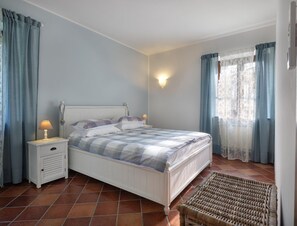 Ein Schlafzimmer mit einem Bett, Nachttischen, einer Lampe, einem Korbstuhl und einem Fenster mit Vorhängen.