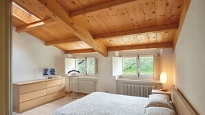 Une chambre avec un plafond en bois, un lit avec une literie à motifs, une commode, un téléviseur et deux fenêtres donnant sur un paysage verdoyant.