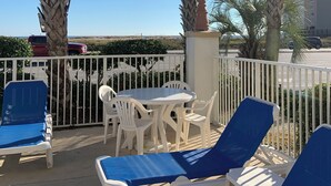 A patio area with white plastic chairs and tables, palm trees, and a view of the road and buildings in the background. - BEAUTIFUL GULF VIEW 2B/2B BALCONY OVER THE POOL,EASY BEACH ACCESS (Orange Beach)