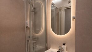 Une salle de bain moderne dotée d’une douche à parois de verre, d’un lavabo blanc avec un meuble-vasque en bois et d’un miroir éclairé par un luminaire placé sous le meuble.