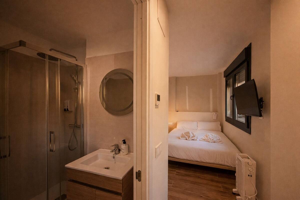 A bedroom with a bed, a bedside table, a TV, and a bathroom with a shower, sink, and mirror.