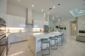 A modern kitchen with a central island, bar stools, and a dining area with a sofa. - Villa Aesthetic View - great view, on water, pool, spa, gulf access (Cape Coral)