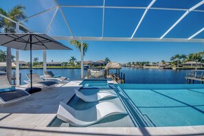 An indoor swimming pool with lounge chairs and umbrellas, a view of a marina with boats, and a clear blue sky. - Villa Aesthetic View - great view, on water, pool, spa, gulf access (Cape Coral)