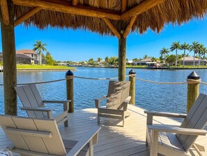 A wooden deck with chairs and a thatched roof, overlooking a body of water with palm trees and buildings in the background. - Villa Aesthetic View - great view, on water, pool, spa, gulf access (Cape Coral)