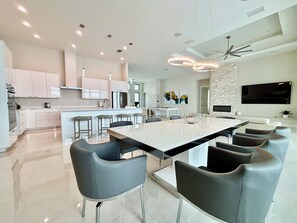 A modern kitchen with white cabinets, a central island with bar stools, a dining area with a large table and chairs, a flat-screen TV mounted on the wall, and a ceiling fan. - Villa Aesthetic View - great view, on water, pool, spa, gulf access (Cape Coral)