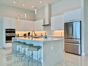 A modern kitchen with stainless steel appliances, a central island with bar stools, and pendant lighting. - Villa Aesthetic View - great view, on water, pool, spa, gulf access (Cape Coral)