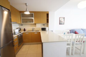 A modern kitchen with wooden cabinets, stainless steel appliances, a white countertop, and a dining area with white chairs. - Panoramic 24 by Parc Mont-roig Apartment (Miami Playa)