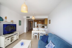Ein Wohnzimmer mit Fernseher, weißem Unterhaltungsschrank, blauem Sofa und einem Essbereich mit weißem Tisch und Stühlen.