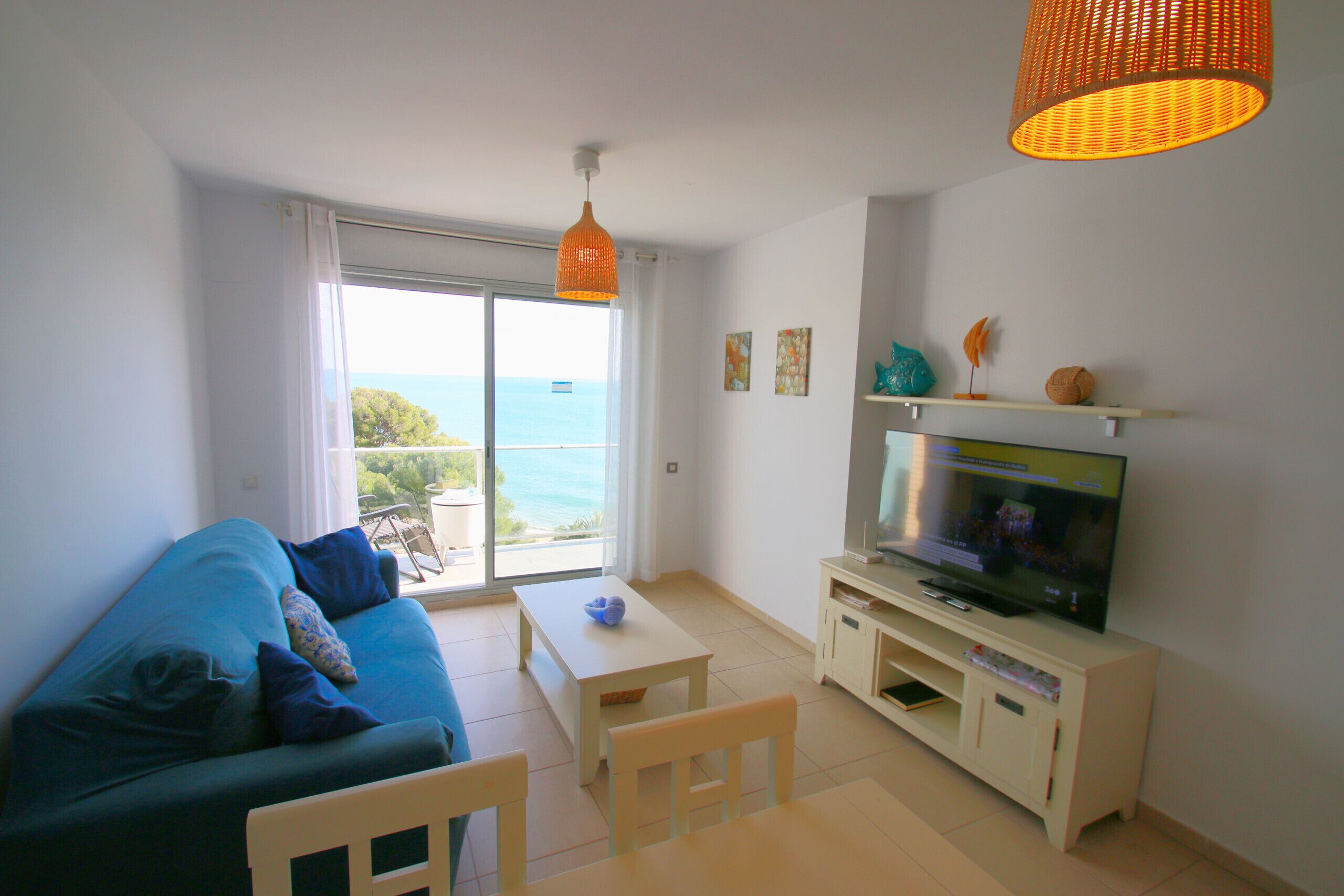 A living room with a sofa, a TV, a table, and a balcony with a view of the sea.