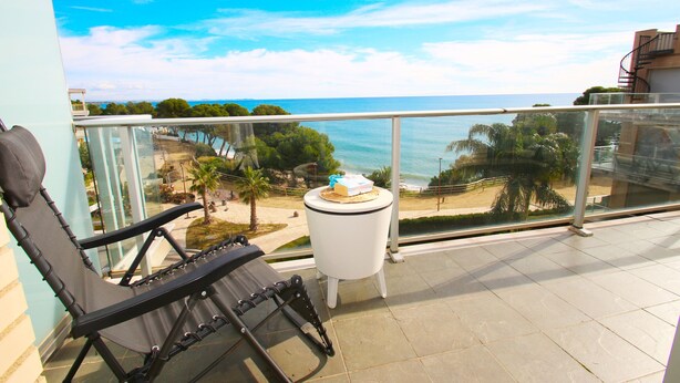 En balkon med en sort loungeseng, et lille rundt bord med kage, og udsigt til stranden og havet.