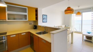 Una cocina moderna con armarios de madera, un lavamanos de acero inoxidable y encimera blanca. Hay un comedor con mesa y sillas, y vistas al mar a través de amplios ventanales.