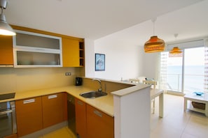 Eine moderne Küche mit Holzschränken, einer Edelstahlspüle und einer weißen Arbeitsplatte. Ein Essbereich mit Tisch und Stühlen lädt ein, und große Fenster bieten Blick auf das Meer.