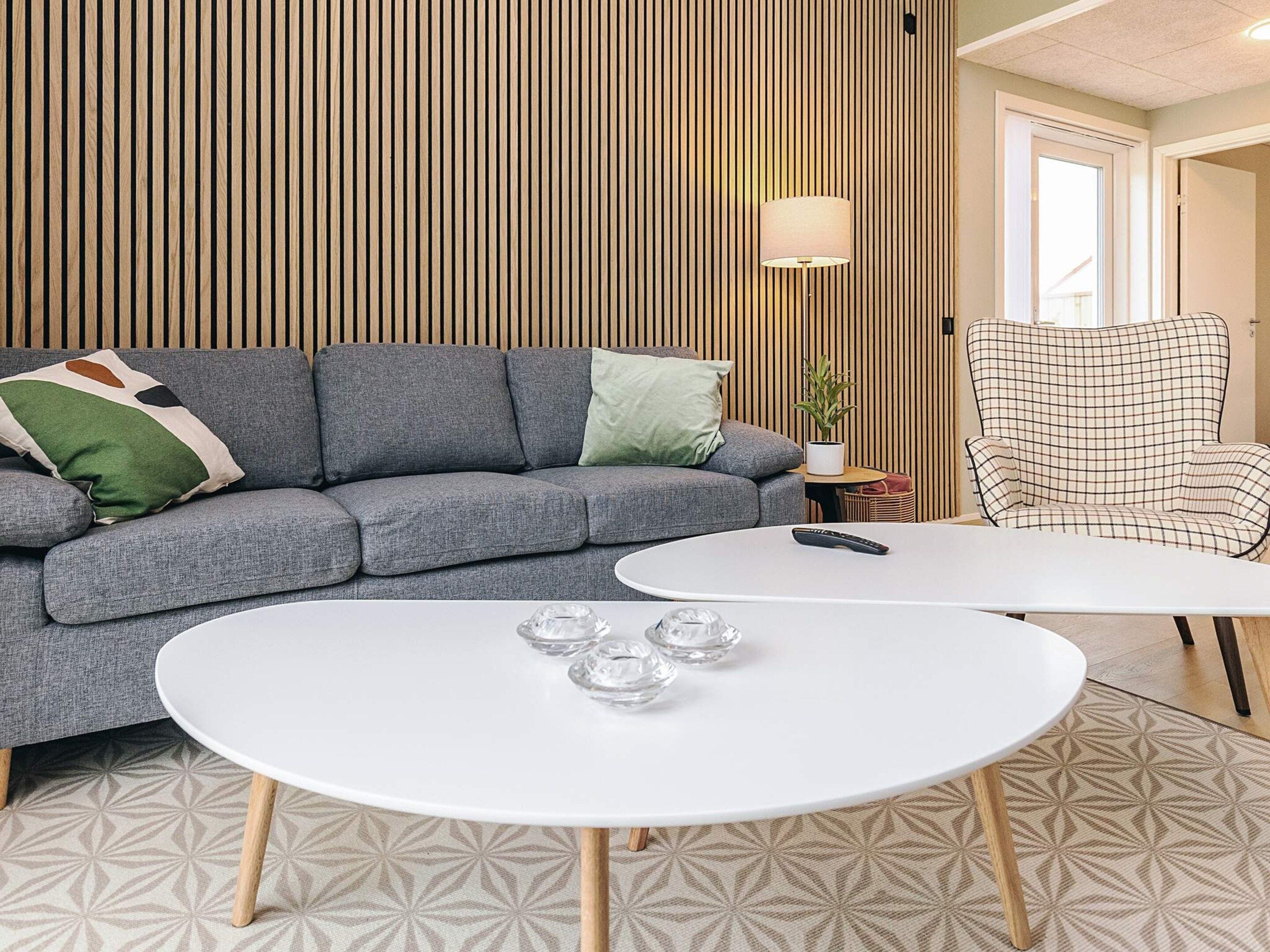 A modern living room with a grey sofa, a white coffee table with glass objects, a plaid armchair, and a wooden slatted wall.