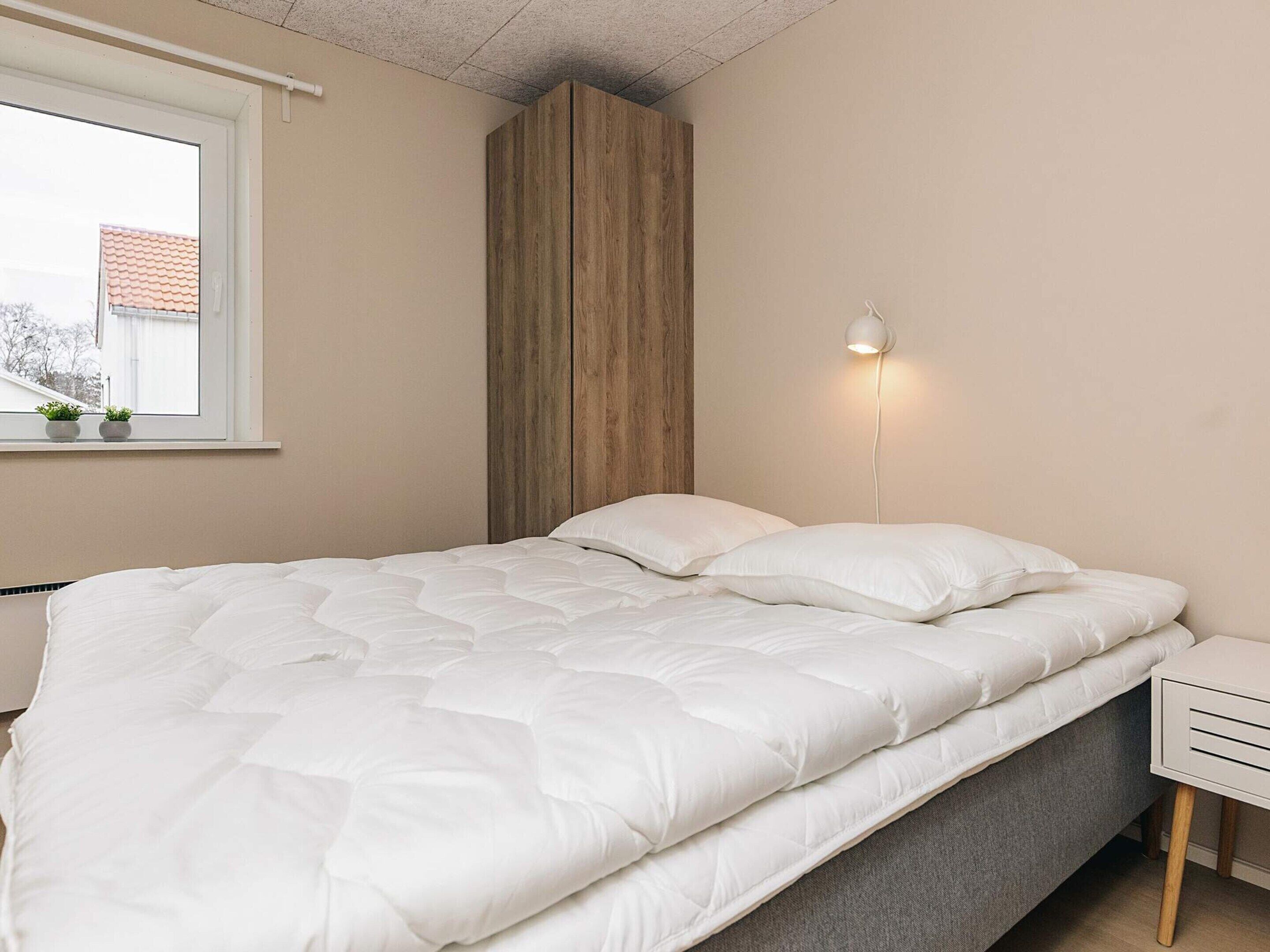 A bed with white bedding, a bedside table, a wall-mounted light, and a window with a view of a building.
