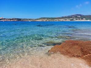 A coastal beach with clear shallow waters, a rocky foreground, and a distant hill with buildings. - House with sea view, 2 minutes walk to the beach in Residence with Pool. (Sardegna)