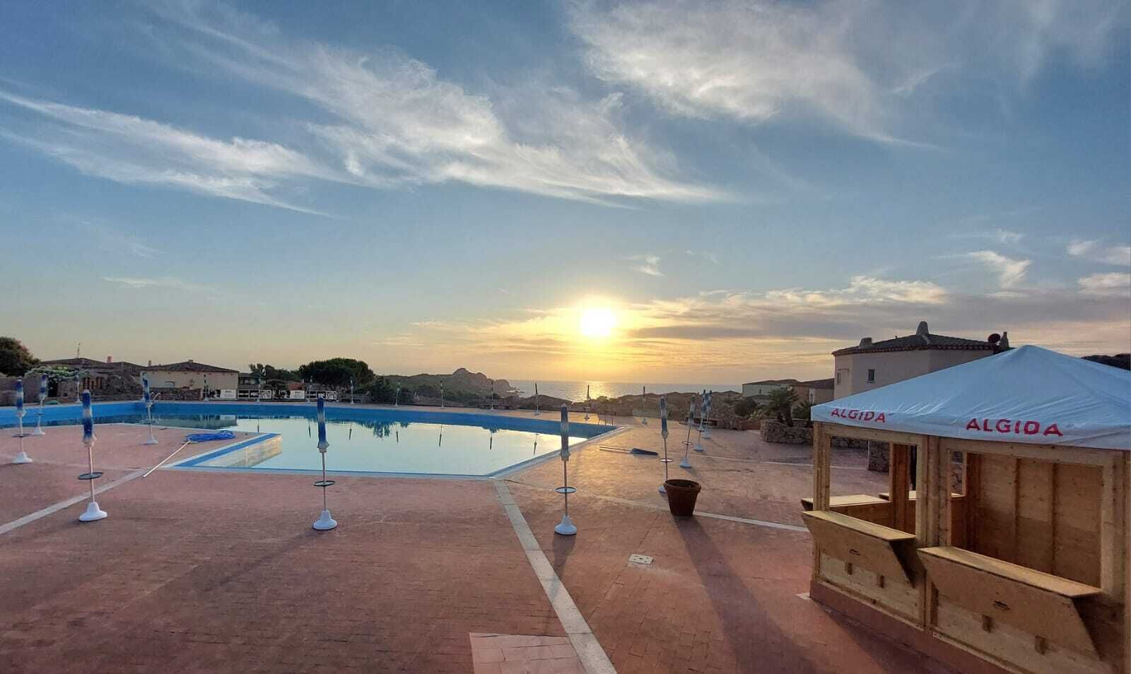 Una piscina con vista sul tramonto, una chiosca in legno e una tenda con la scritta 'ALGIDA'.