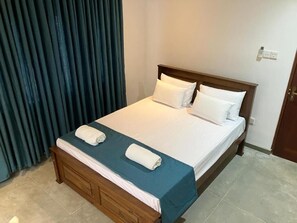 A neatly made bed with white linens and a blue bedspread, a wooden headboard, and a dark curtain to the left. - Seth Villa (Matara)