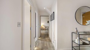A hallway with a wooden floor, a round mirror on the wall, and a black metal shelf with books and decorative items. - 5 Mins Walk to Battersea Park Battersea Retreat (London)