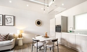 Eine moderne Küche mit weißen Schränken, einem runden Esstisch mit vier Stühlen, einem kleinen Beistelltisch mit Lampe und einer dekorativen Vase.