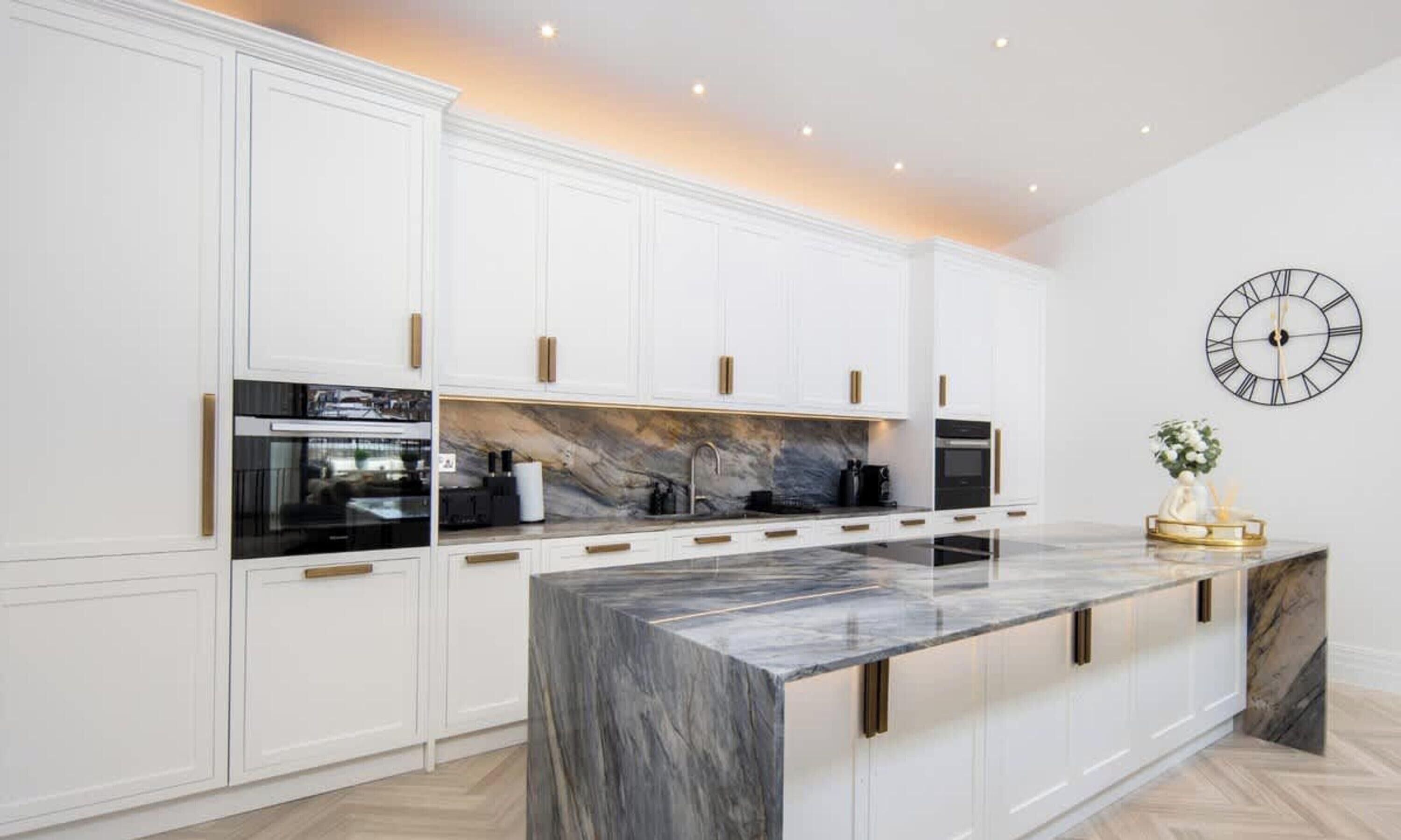 A modern kitchen with white cabinetry, a marble countertop, and a black clock on the wall.