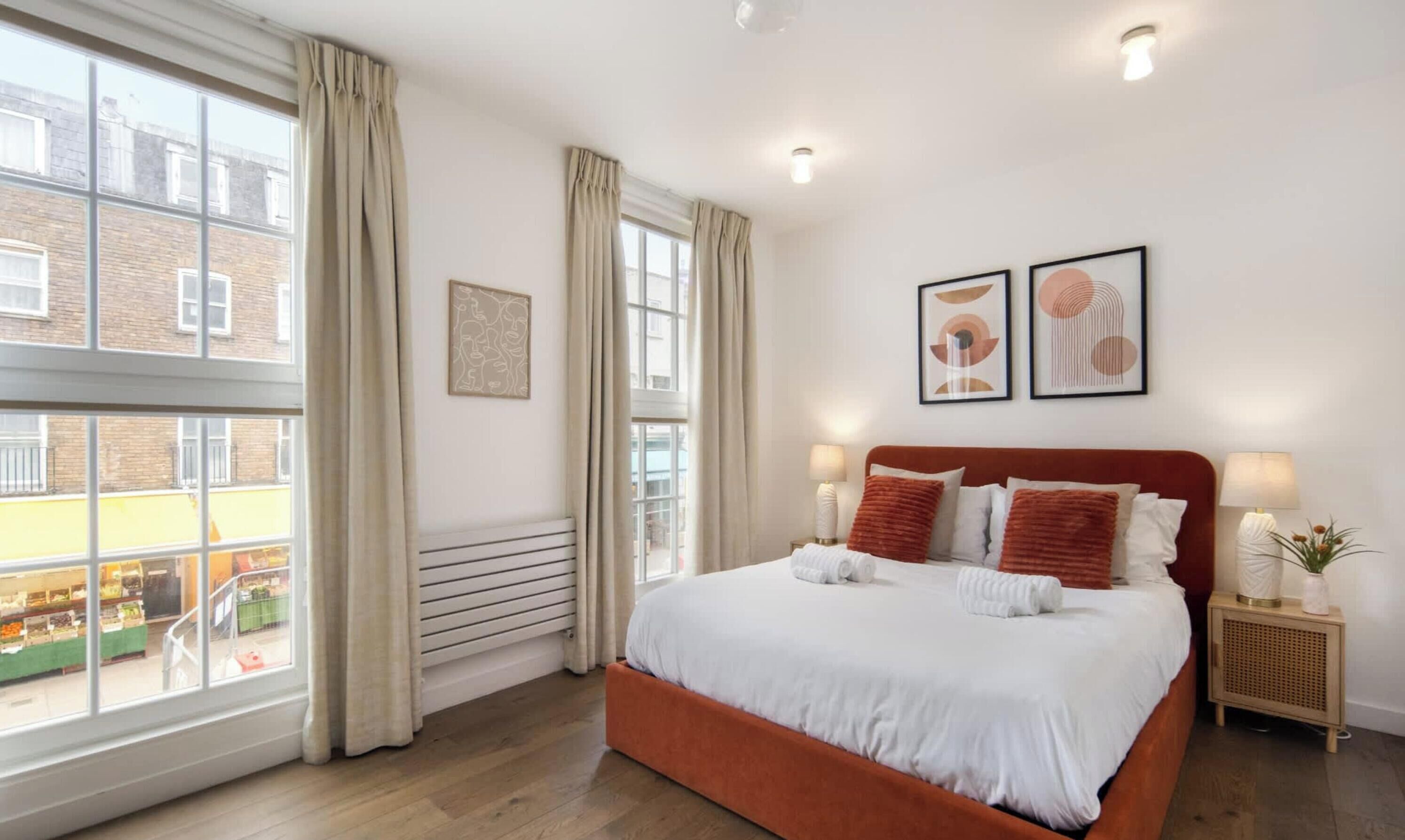 A bedroom with a large window, a bed with white linens, and two framed artworks on the wall.