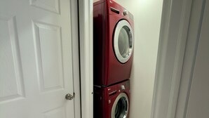 Uma lavanderia com máquina de lavar e secadora empilhadas, uma porta branca e piso de azulejos bege.