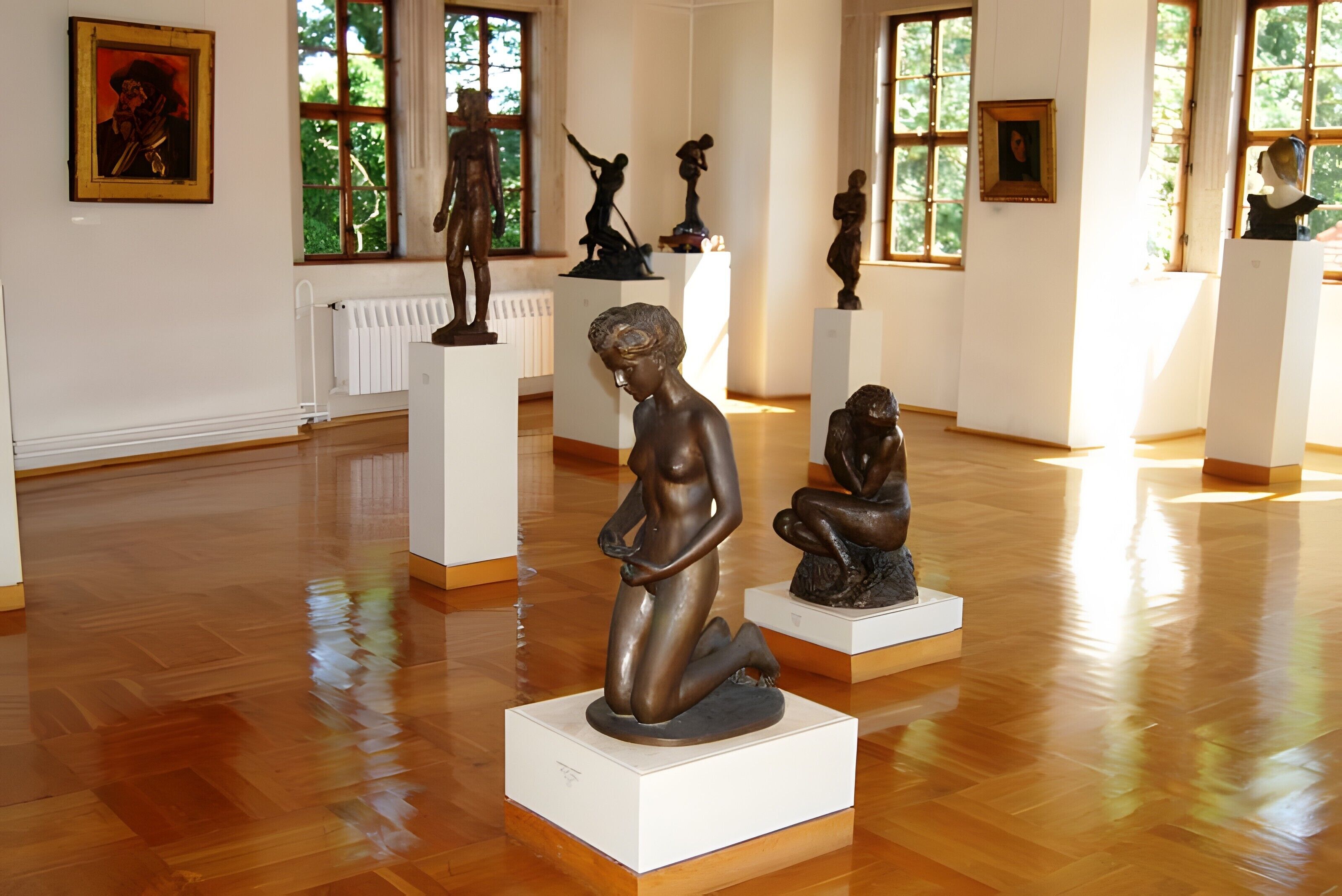 Una galería de arte con esculturas sobre pedestales, pisos de madera y ventanas amplias.
