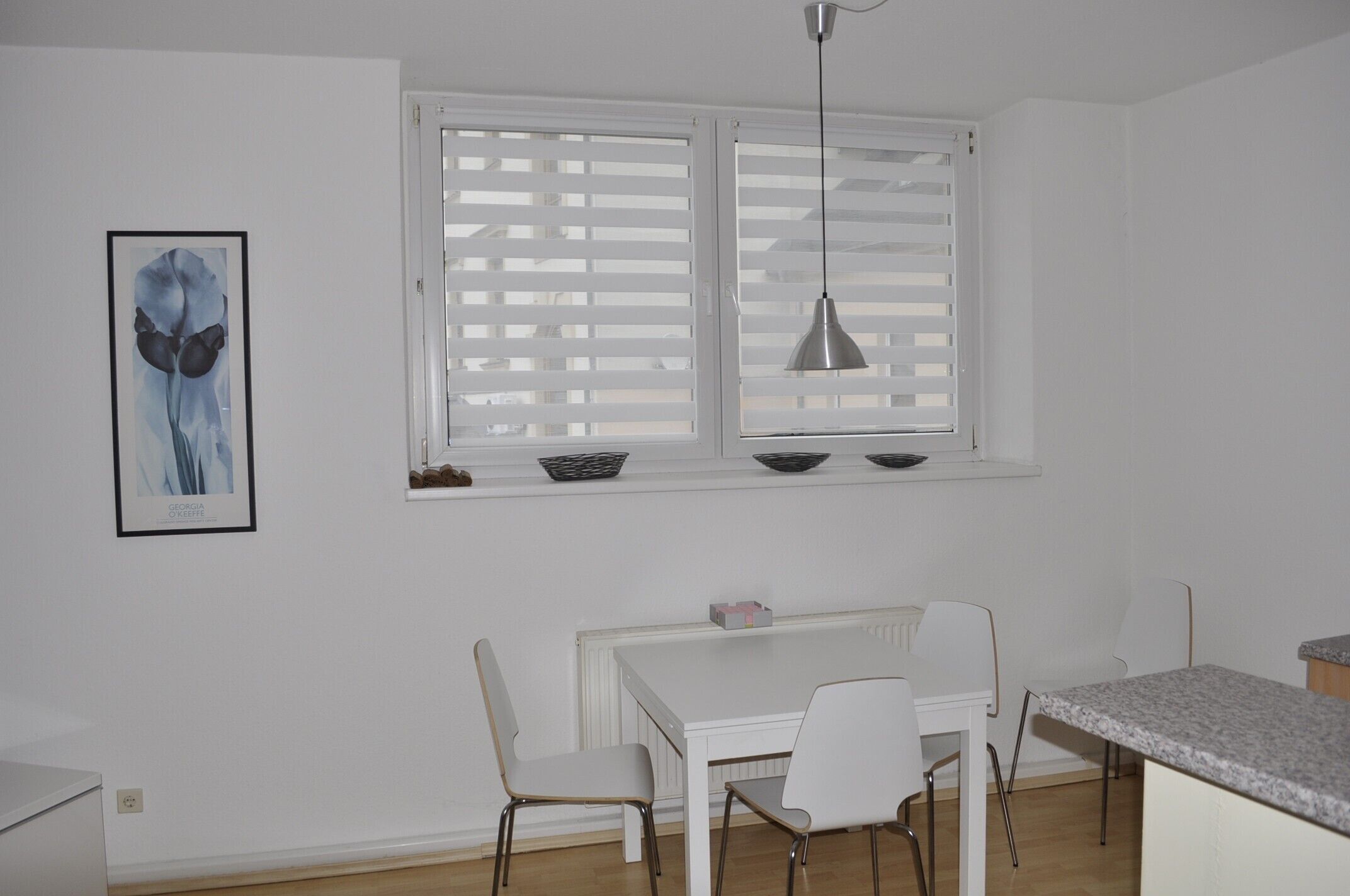 Ein weißes Esszimmer mit Tisch, Stühlen, einem Fenster mit Jalousien und einer Pendelleuchte.