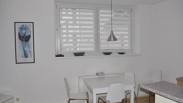Ein weißes Esszimmer mit Tisch, Stühlen, einem Fenster mit Jalousien und einer Pendelleuchte.