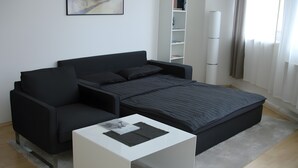 Une chambre moderne avec un canapé noir, une table basse blanche et un lit doté d’un matelas gris.
