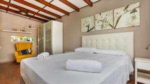 Una habitación de hotel con cama, un sillón amarillo, cabecera blanca y obras de arte en la pared.
