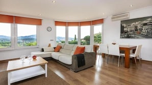 Un salón moderno con sofá, mesa de comedor y sillas, ventanas amplias con vistas, y un reloj en la pared.