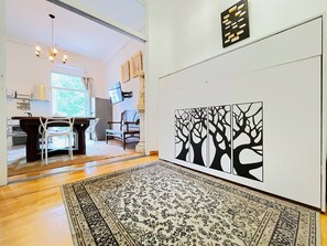Ein Zimmer mit Holzboden, einem gemusterten Teppich, einem Essbereich mit Stühlen und einem Tisch sowie einer Wand mit einer Schwarz-Weiß-Baum-Kunst.'
