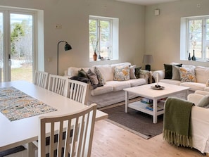 Un salon moderne avec un ensemble de canapés blancs, une table à manger et une table basse. Deux fenêtres laissent entrer la lumière naturelle et offrent une vue sur l’extérieur.