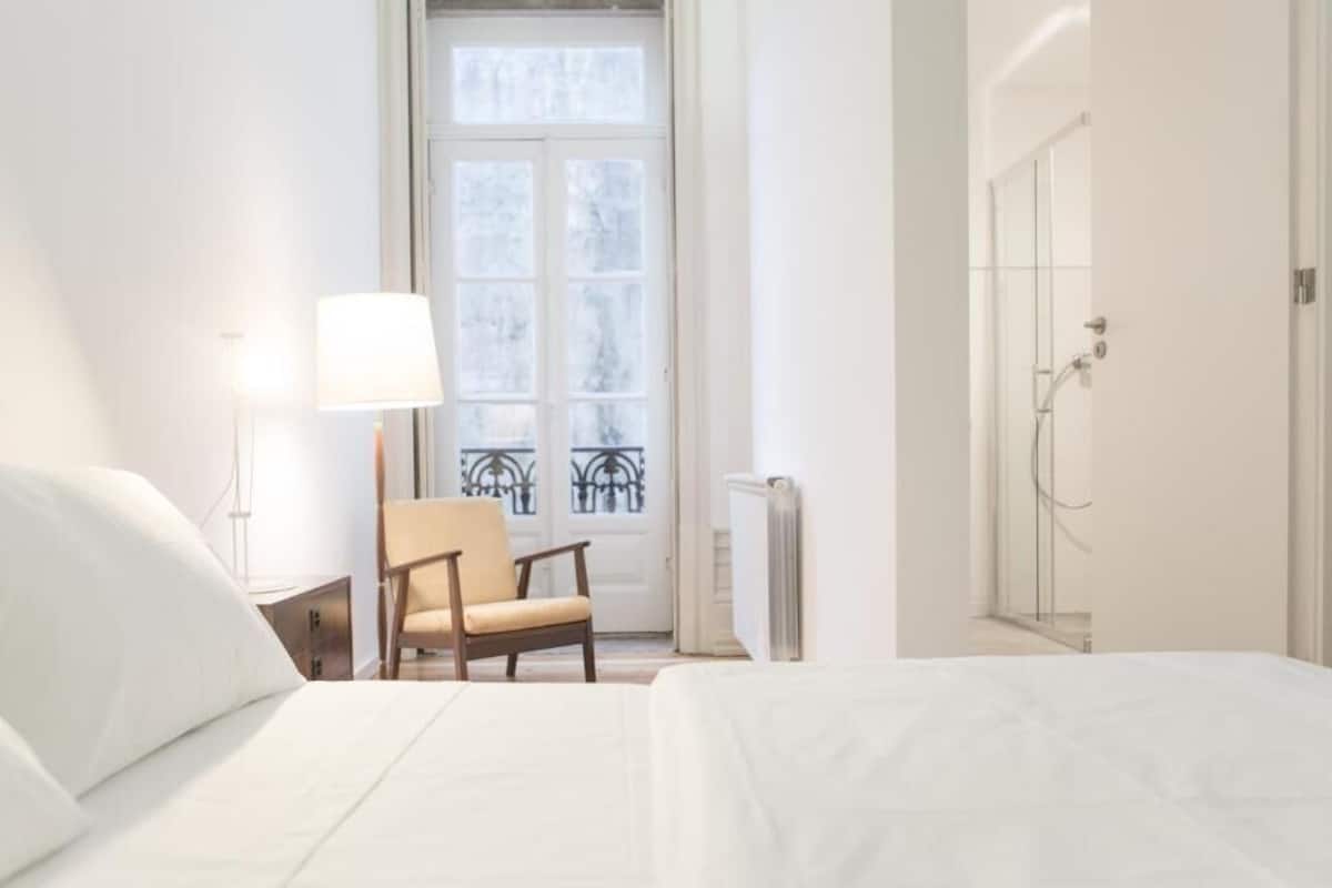 A hotel room with a bed, a chair, a lamp, a radiator, and a door leading to a shower.