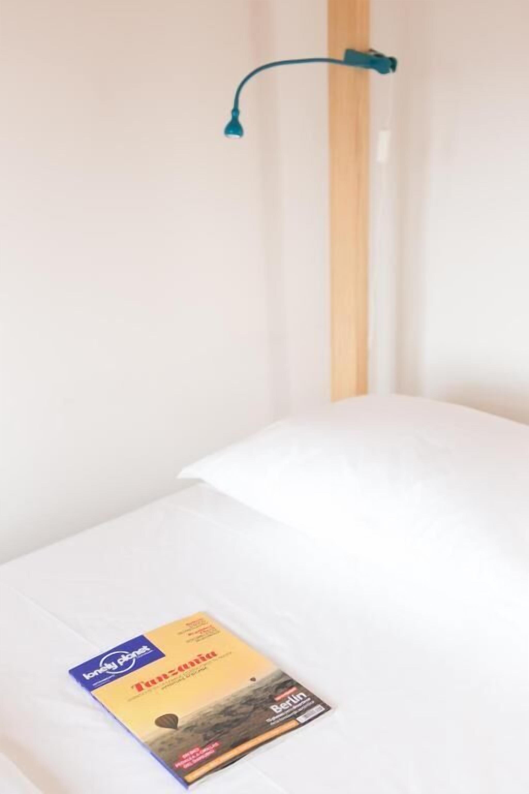 A single bed with a white mattress and a single pillow, a teal desk lamp, and a Lonely Planet travel guide on the bed.