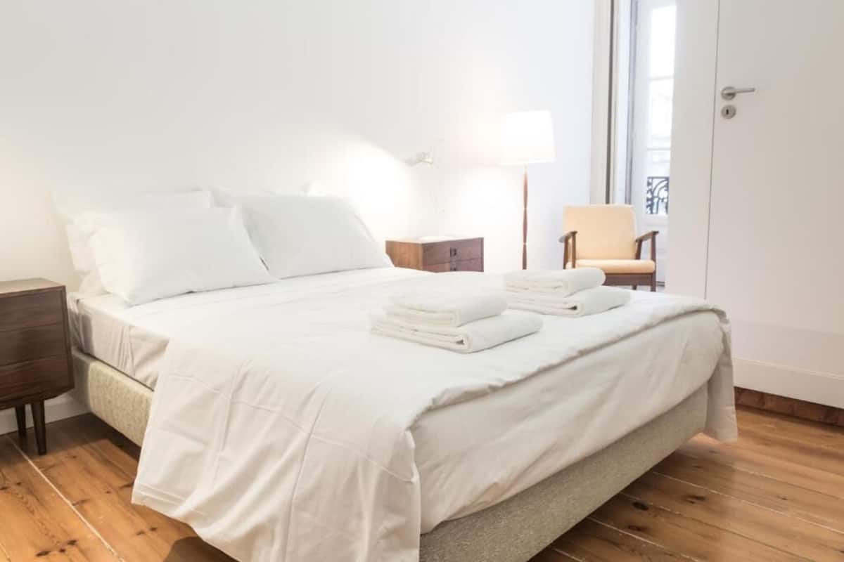 A neatly made bed with white linens, a bedside table, a chair, and a lamp.