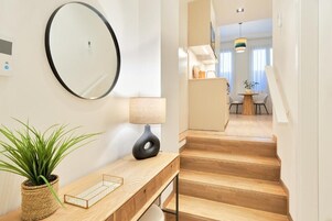 Un interior moderno con un escritorio de madera, una planta en maceta, un espejo redondo y una escalera que lleva a un comedor.