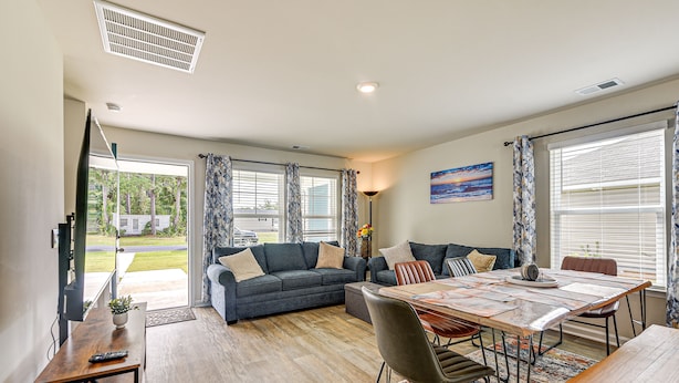A spacious living area with a sofa, dining table, and chairs. There is a large window with curtains, a painting on the wall, and a door leading outside. - Ocean Breeze Escape! (Ocean Isle Beach)