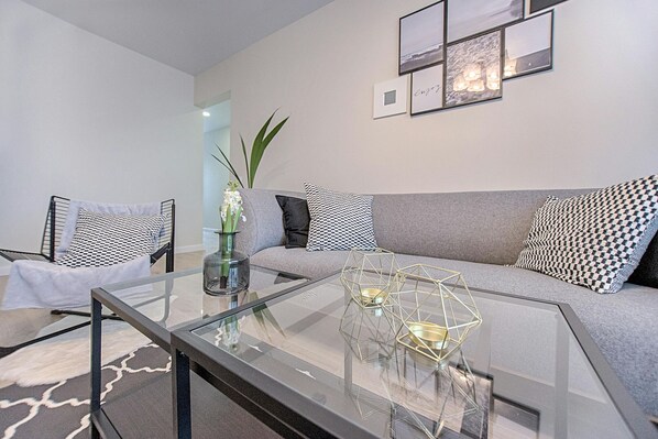 Ein modernes Wohnzimmer mit einem Glastisch, einem grauen Sofa, gemusterten Kissen und gerahmten Bildern an der Wand.