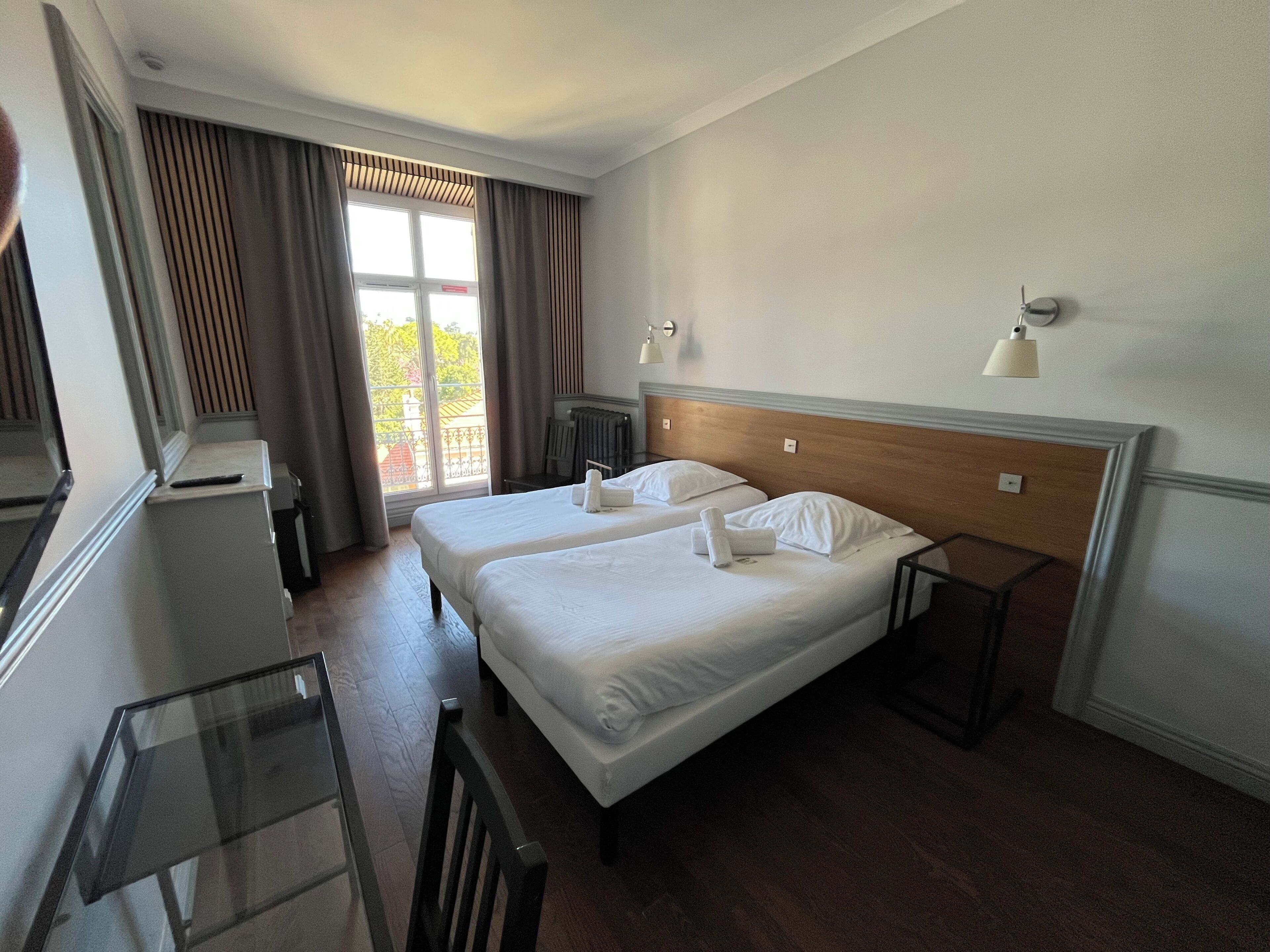 Une chambre d’hôtel avec deux lits, un bureau, une chaise, un radiateur et une fenêtre avec des rideaux.