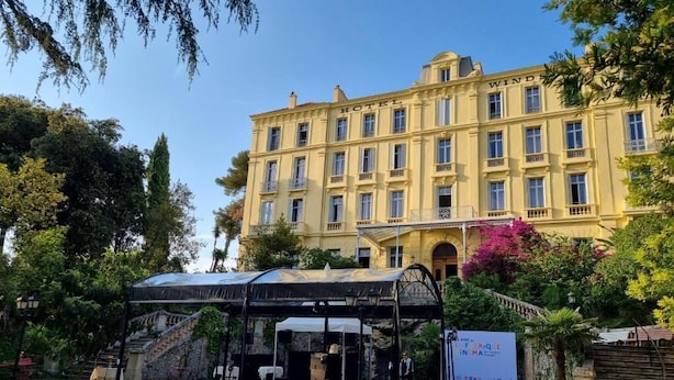 Un hôtel à la façade classique, doté d’un espace extérieur couvert avec des tables, et d’une enseigne indiquant un événement.