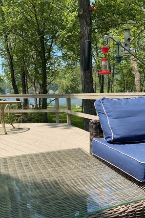 Een terras met een rieten bank en een glazen tafel, met uitzicht op een meer met bomen en een balustrade.