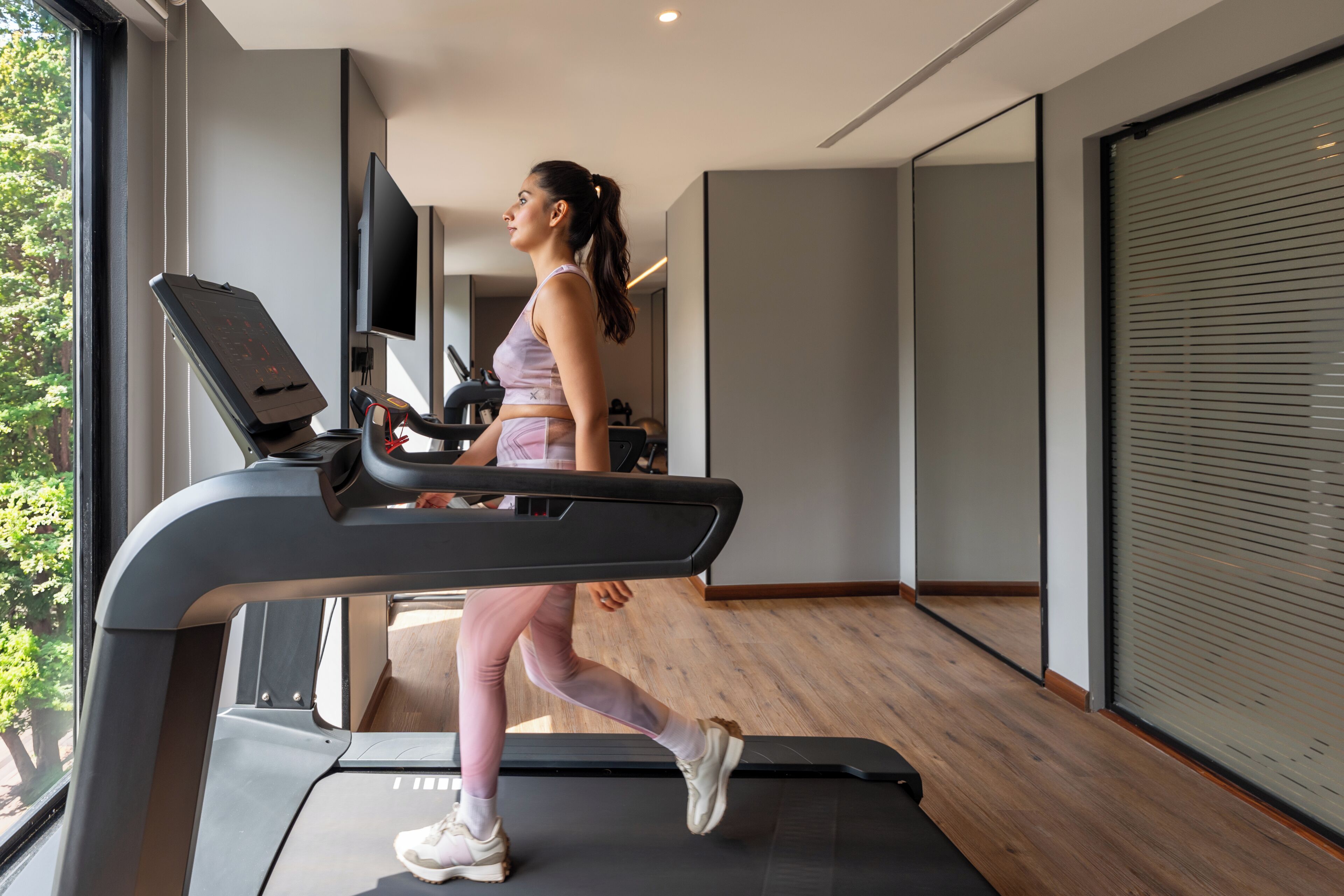 Uma mulher corre na passadeira de um ginásio moderno, com uma janela grande, uma televisão de ecrã plano e uma porta de correr.