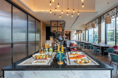 A buffet setup with various food items and beverages, a kitchen area in the background, and a dining area with tables and chairs.. Park Inn By Radisson Jabalpur