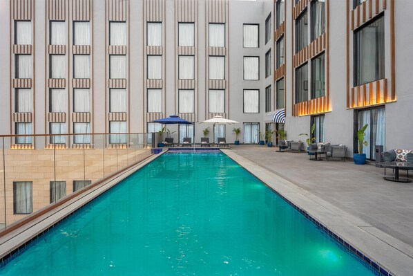 A swimming pool with a tiled edge, surrounded by lounge chairs and umbrellas, and a building with large windows and vertical wooden panels. - Park Inn By Radisson Jabalpur (Jabalpur)