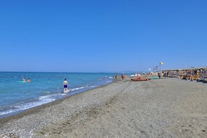 Ein Kiesstrand mit badenden und spazierenden Personen, einem Rettungsturm und Liegestühlen.