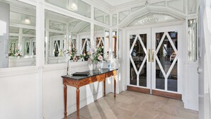 A grand entrance with a glass door, a white wall with a mirror, and a wooden console table with a vase of flowers. - The Degas Sea View Suite (Torquay)