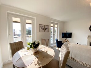 A dining area with a round table, chairs, and a potted plant. There is a sofa and a television in the background. - The Degas Sea View Suite (Torquay)