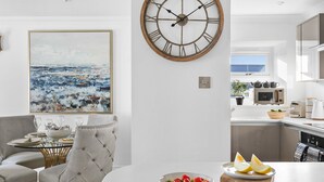 A modern kitchen with a large wall clock, a dining area with a round table and chairs, and a view of the sea through the window. - The Renoir Sea View Suite (Torquay)